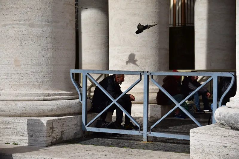 A pigeon flies between pale stone columns while people rest in the shade behind a metal barrier